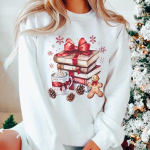 May include: White sweatshirt with a festive graphic. The design features a stack of books tied with a red bow, a mug of whipped cream, a gingerbread man, pine cones, and snowflakes. A cosy winter theme.