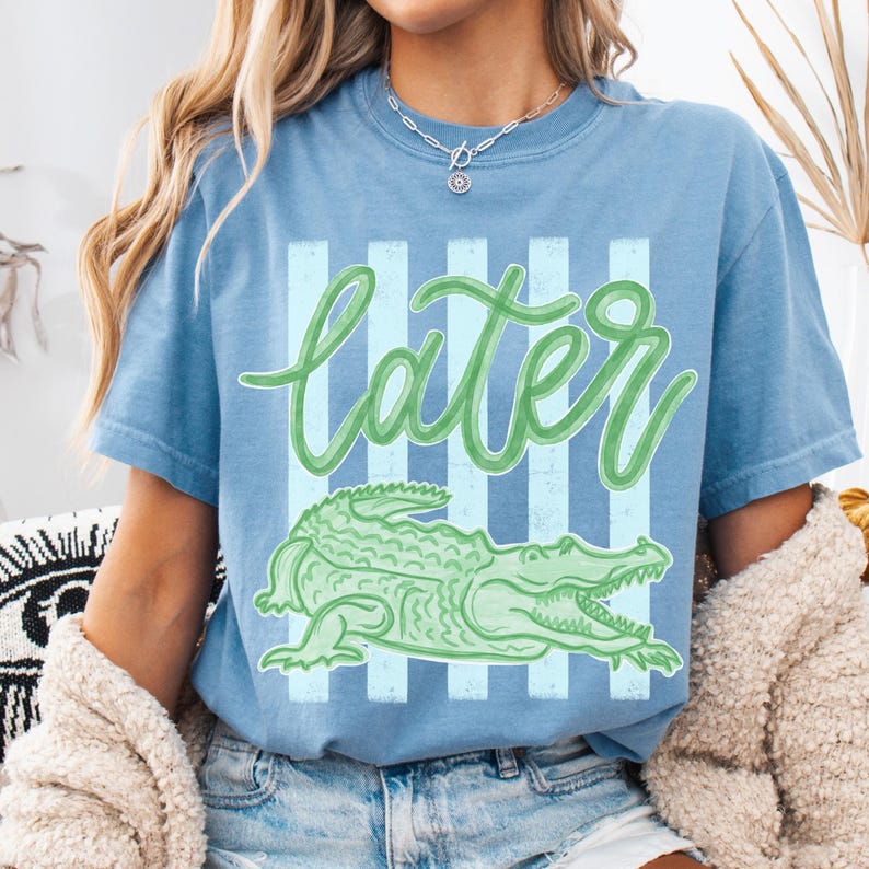 Puede incluir: Camiseta azul claro con un gr&aacute;fico de cocodrilo verde y la palabra "Later" en fuente cursiva. El dise&ntilde;o incluye rayas verticales de color azul claro. La camiseta es de estilo casual.