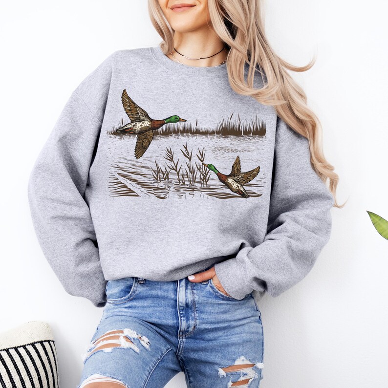 K&ouml;nnte beinhalten: Ein hellgraues Sweatshirt mit einer detaillierten Illustration von zwei Enten im Flug &uuml;ber Wasser mit Schilf. Die Enten haben braune, gr&uuml;ne und wei&szlig;e Markierungen. Das Sweatshirt ist aus weichem Material.