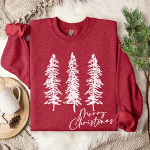 May include: A red sweatshirt with a crew neck and long sleeves. The front features a white graphic of three stylized Christmas trees and the words "Merry Christmas" in a script font. The sweatshirt is displayed on a white fur-like surface.