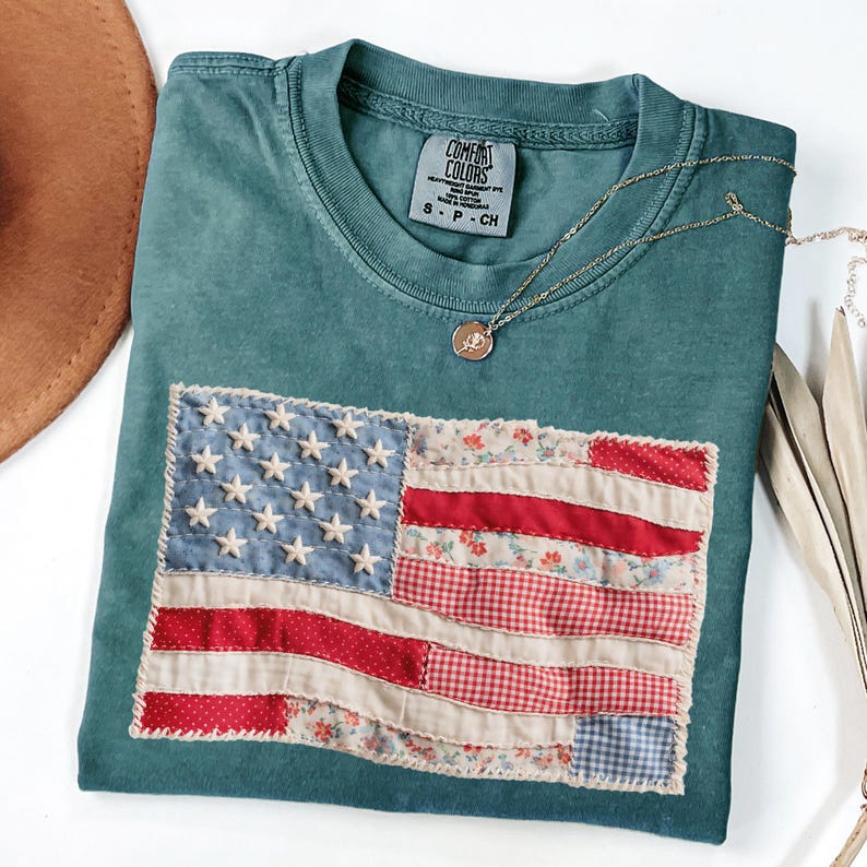 K&ouml;nnte beinhalten: Ein teal Comfort Colors T-Shirt mit einem aufgen&auml;hten amerikanischen Flaggen-Design. Die Flagge zeigt einen Patchwork aus blauem Denim mit wei&szlig;en Sternen, roten und floral gemusterten Streifen. Eine goldene Halskette mit Anh&auml;nger liegt auf dem Shirt.