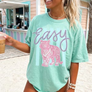 K&ouml;nnte beinhalten: Hellgr&uuml;nes T-Shirt mit dem Wort "Easy" in Schreibschrift &uuml;ber einer rosa Tiger-Illustration. Der Tiger ist vor einem hellblau gestreiften Hintergrund platziert. Das Shirt hat eine lockere Passform.
