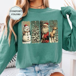 May include: Teal-colored sweatshirt featuring a festive graphic. The graphic includes a snowman, Christmas tree, Santa Claus, and a reindeer, all within rectangular frames. The text "COMFORT COLORS" is in a white circular badge.