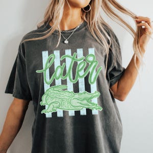 Puede incluir: Camiseta gris oscuro con un dise&ntilde;o de cocodrilo verde y la palabra "later" en letra cursiva. El dise&ntilde;o incluye rayas verticales de color azul claro. Un estilo casual y unisex.