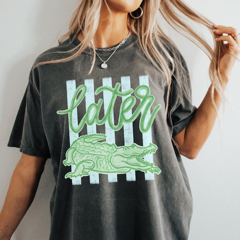 Puede incluir: Camiseta gris oscuro con un dise&ntilde;o de cocodrilo verde y la palabra "later" en letra cursiva. El dise&ntilde;o incluye rayas verticales de color azul claro. Un estilo casual y unisex.