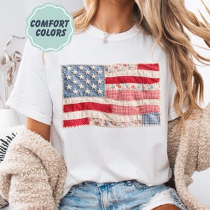 K&ouml;nnte beinhalten: Wei&szlig;es T-Shirt mit einem Patchwork-Design der amerikanischen Flagge. Die Flagge verwendet blaue, rote und wei&szlig;e Stoffe mit Stern- und Streifendetails. Der Text "COMFORT COLORS" befindet sich in einem hellgr&uuml;nen Kreis.