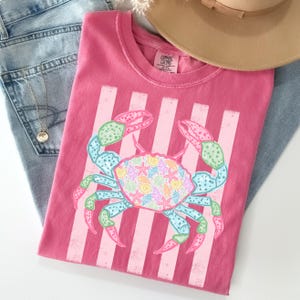 May include: A pink t-shirt featuring a crab design with a striped background. The crab is decorated with seashells and starfish in pastel colors. The shirt is folded on top of a pair of blue jeans and a tan hat.