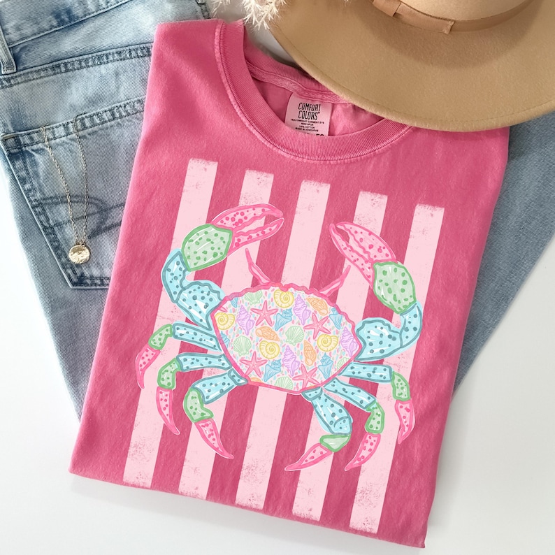 May include: A pink t-shirt featuring a crab design with a striped background. The crab is decorated with seashells and starfish in pastel colors. The shirt is folded on top of a pair of blue jeans and a tan hat.