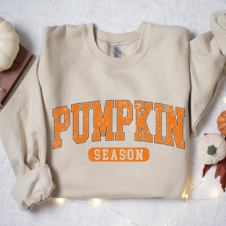 May include: A beige sweatshirt with the words "PUMPKIN SEASON" in orange block letters. The text has a distressed, vintage look. The sweatshirt is folded, with the sleeves visible. The overall theme is autumn and fall.
