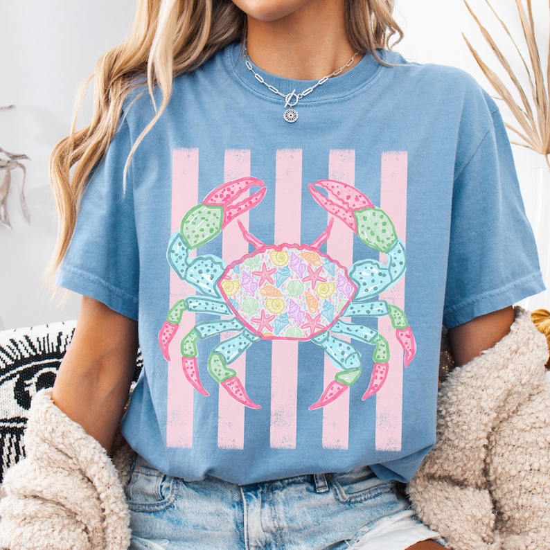 May include: A light blue t-shirt featuring a colorful crab graphic. The crab has pink and green claws, and a body filled with seashells and starfish. Pink vertical stripes are behind the crab.