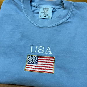 May include: A light blue t-shirt with the word "USA" embroidered in white above an embroidered American flag. The shirt has a crew neck and the brand "Comfort Colors" is printed on the tag.