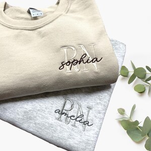 Puede incluir: Dos sudaderas con las letras "RN" y un nombre bordado. La sudadera de arriba es beige con el nombre "sophia" bordado en negro. La sudadera de abajo es gris con el nombre "amelia" bordado en negro.