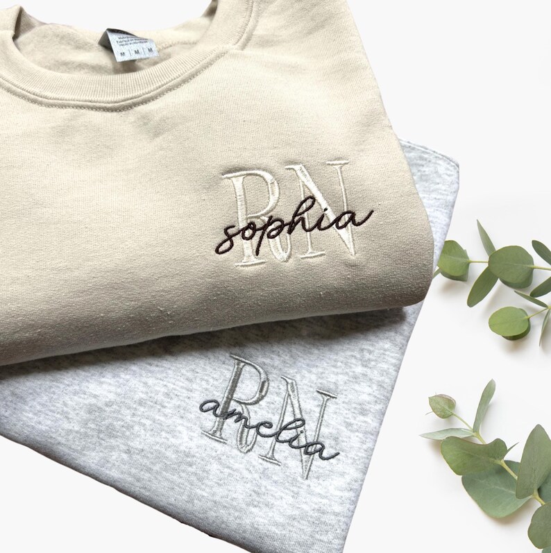 Puede incluir: Dos sudaderas con las letras "RN" y un nombre bordado. La sudadera de arriba es beige con el nombre "sophia" bordado en negro. La sudadera de abajo es gris con el nombre "amelia" bordado en negro.