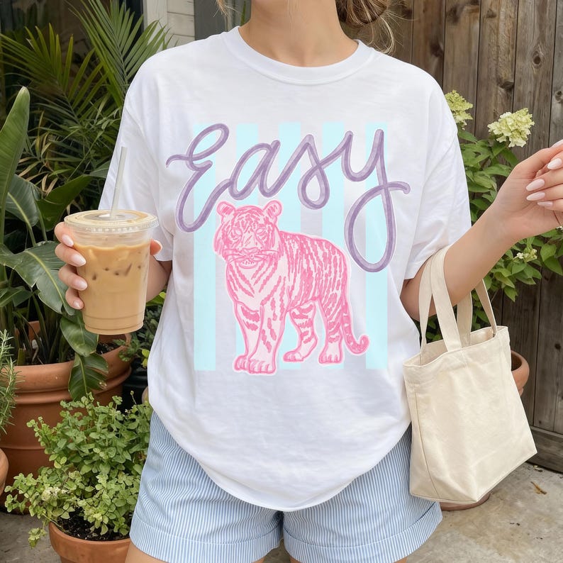 K&ouml;nnte beinhalten: Wei&szlig;es T-Shirt mit einem rosa Tiger-Motiv und dem Wort "Easy" in lavendelfarbener Schrift. Der Tiger ist vor einem hellblau gestreiften Hintergrund platziert. Das Shirt wird mit blau-wei&szlig; gestreiften Shorts getragen.