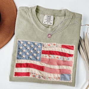 K&ouml;nnte beinhalten: Olivgr&uuml;nes T-Shirt mit einem Patchwork-Design der amerikanischen Flagge. Die Flagge zeigt blauen Stoff mit wei&szlig;en Sternen und rot- und blumenmusternden Streifen. Das Shirt hat ein Comfort Colors-Etikett und eine silberne Halskette.