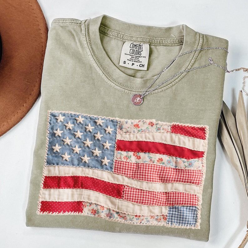 K&ouml;nnte beinhalten: Olivgr&uuml;nes T-Shirt mit einem Patchwork-Design der amerikanischen Flagge. Die Flagge zeigt blauen Stoff mit wei&szlig;en Sternen und rot- und blumenmusternden Streifen. Das Shirt hat ein Comfort Colors-Etikett und eine silberne Halskette.