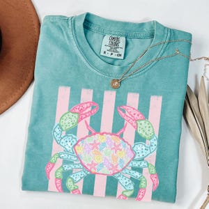 May include: Teal Comfort Colors t-shirt featuring a crab design with pink and pastel accents. The crab is centered on the shirt, with pink vertical stripes behind it. A gold necklace with a pendant is draped across the shirt.