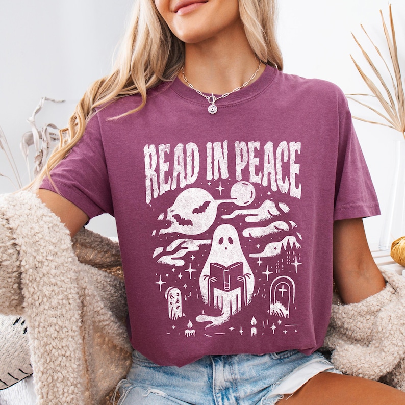 May include: A maroon t-shirt with the text "READ IN PEACE" in white, featuring a graphic of a ghost reading a book, with bats, a moon, and stars. The design has a spooky, Halloween theme.