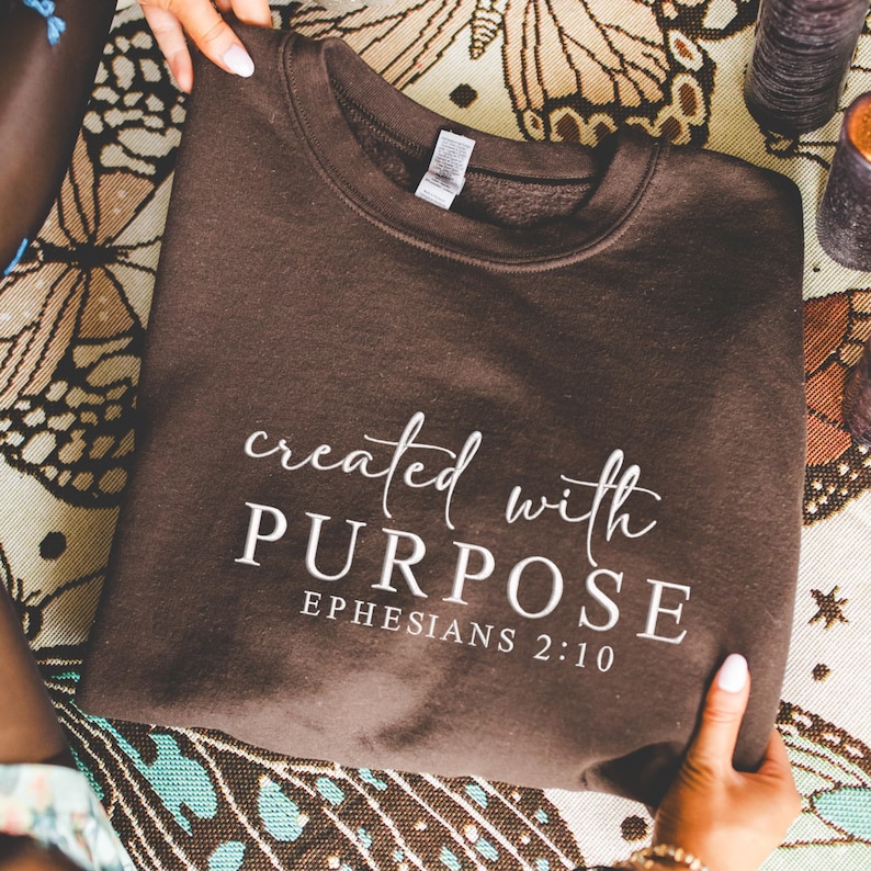 May include: A dark brown sweatshirt with the words "created with PURPOSE" and "EPHESIANS 2:10" printed in white script. The sweatshirt is displayed on a patterned surface.