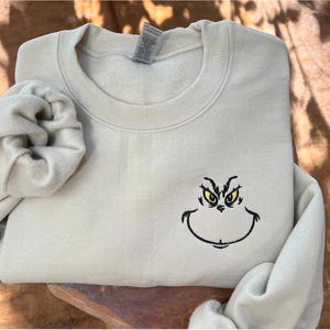 Puede incluir: Una sudadera beige claro con cuello redondo y mangas largas. La sudadera presenta una cara de Grinch bordada con ojos amarillos y un contorno negro. Una scrunchie a juego est&aacute; al lado de la sudadera.