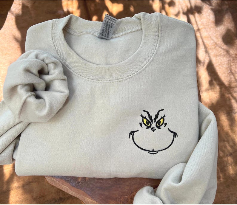 Puede incluir: Una sudadera beige claro con cuello redondo y mangas largas. La sudadera presenta una cara de Grinch bordada con ojos amarillos y un contorno negro. Una scrunchie a juego est&aacute; al lado de la sudadera.