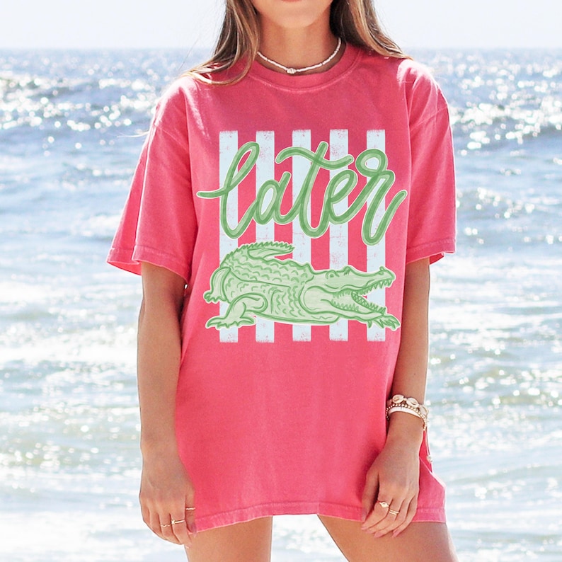 Puede incluir: Camiseta de color coral con un gr&aacute;fico de un caim&aacute;n verde y la palabra "later" en una fuente cursiva. El dise&ntilde;o incluye rayas blancas verticales. La camiseta tiene mangas cortas y un corte holgado.