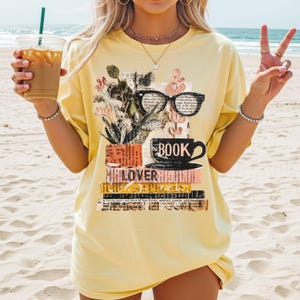 May include: Yellow t-shirt with a graphic design featuring a stack of books, a coffee cup, eyeglasses, and floral elements. The word "BOOK" is printed above the cup, and "LOVER" is printed on the books. The shirt is worn on a beach.