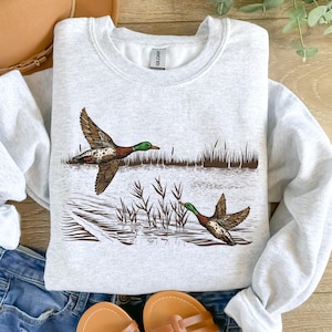 K&ouml;nnte beinhalten: Hellgraues Sweatshirt mit detaillierter Illustration von zwei Enten im Flug &uuml;ber Wasser und Schilf. Die Enten haben braune, gr&uuml;ne und wei&szlig;e Markierungen. Das Design ist mittig auf der Vorderseite platziert.