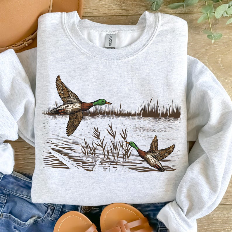 K&ouml;nnte beinhalten: Hellgraues Sweatshirt mit detaillierter Illustration von zwei Enten im Flug &uuml;ber Wasser und Schilf. Die Enten haben braune, gr&uuml;ne und wei&szlig;e Markierungen. Das Design ist mittig auf der Vorderseite platziert.