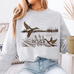 K&ouml;nnte beinhalten: Hellgraues Sweatshirt mit einer detaillierten Illustration von zwei Enten im Flug &uuml;ber Wasser. Die Enten haben braune und wei&szlig; gemusterte Fl&uuml;gel und gr&uuml;ne K&ouml;pfe. Schilf und Wasser sind ebenfalls dargestellt.