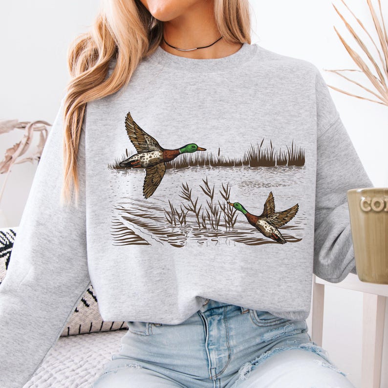 K&ouml;nnte beinhalten: Hellgraues Sweatshirt mit einer detaillierten Illustration von zwei Enten im Flug &uuml;ber Wasser. Die Enten haben braune und wei&szlig; gemusterte Fl&uuml;gel und gr&uuml;ne K&ouml;pfe. Schilf und Wasser sind ebenfalls dargestellt.