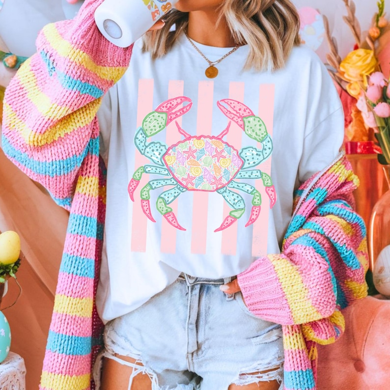 May include: White t-shirt featuring a colorful crab design filled with seashells, starfish, and other sea life. The crab is set against a backdrop of pink vertical stripes. The person is wearing a colorful striped sweater.