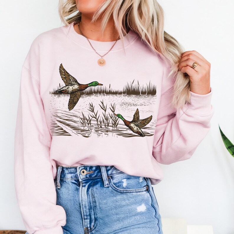K&ouml;nnte beinhalten: Ein hellrosa Sweatshirt mit einer detaillierten Illustration von zwei Enten im Flug &uuml;ber Wasser und Schilf. Die Enten haben braune und gr&uuml;ne Federn. Das Sweatshirt hat lange &Auml;rmel und einen Rundhalsausschnitt.