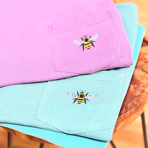 May include: A purple t-shirt and a teal t-shirt stacked on top of each other. Each shirt features a chest pocket with an embroidered bumblebee design. The bees are yellow and black with white wings, positioned centrally on each pocket.
