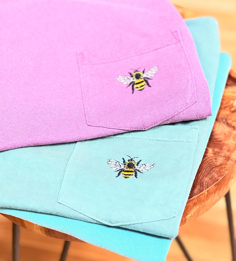 May include: A purple t-shirt and a teal t-shirt stacked on top of each other. Each shirt features a chest pocket with an embroidered bumblebee design. The bees are yellow and black with white wings, positioned centrally on each pocket.