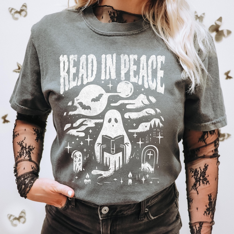 May include: Dark gray t-shirt with a white graphic that reads "READ IN PEACE." The graphic features a ghost reading a book, a full moon, bats, a haunted house, and tombstones. The shirt has short sleeves.