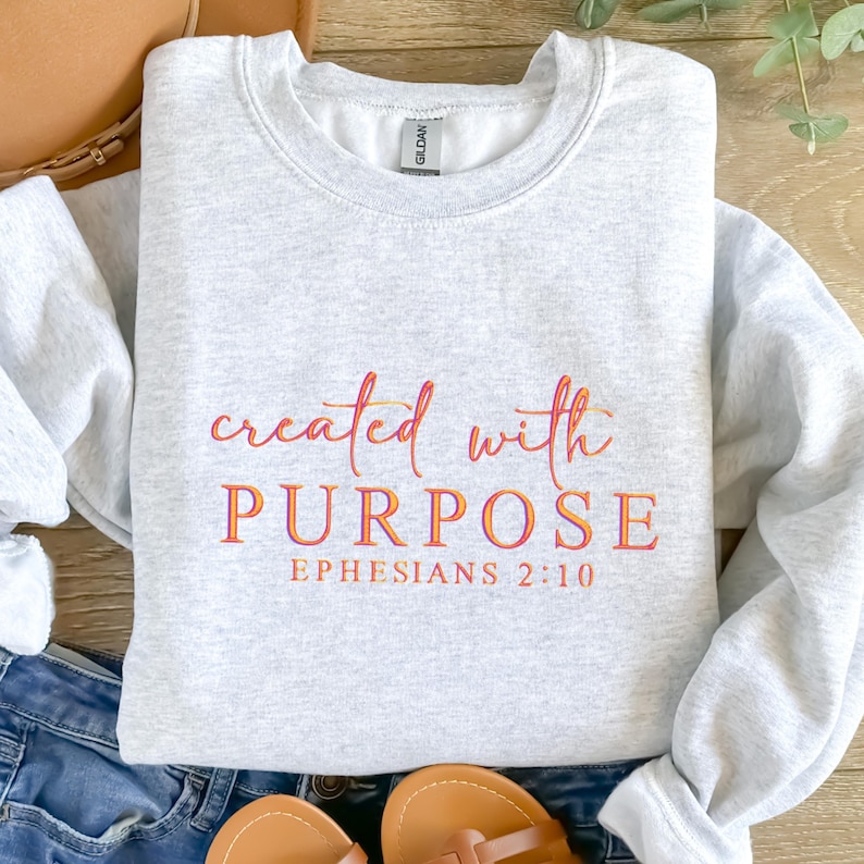 May include: A pale grey sweatshirt featuring the text "created with PURPOSE" in orange and pink script. Beneath, in orange, is "Ephesians 2:10". The sweatshirt is folded on a surface with denim jeans and sandals.