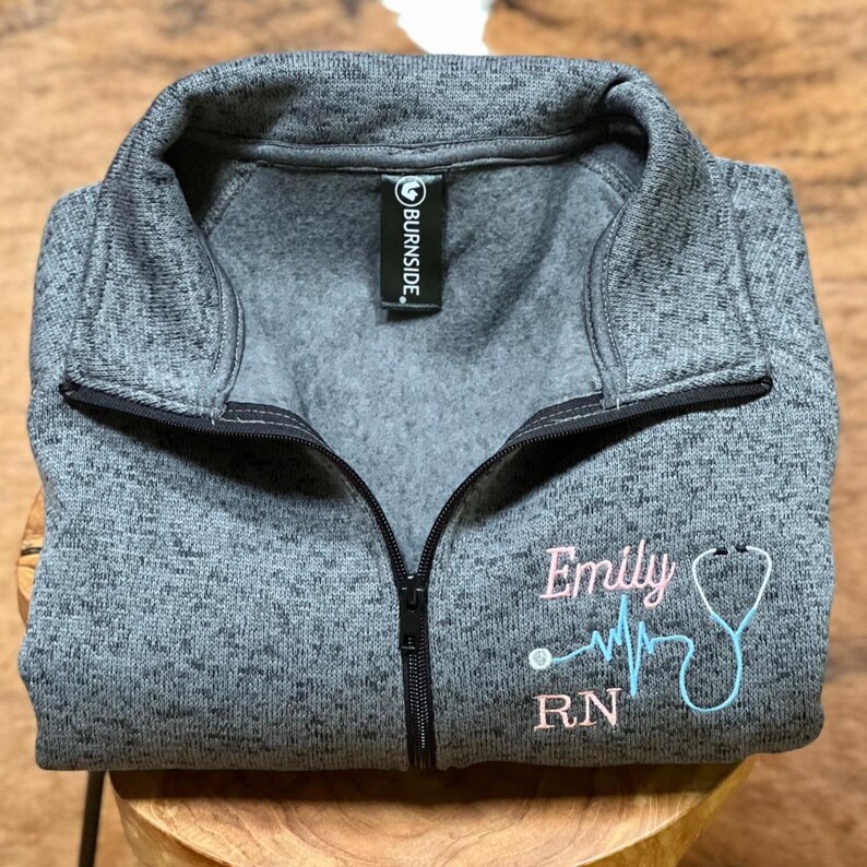 May include: Grey fleece zip-up sweatshirt with a pink embroidered design that says "Emily RN" with a stethoscope and a heart beat line.