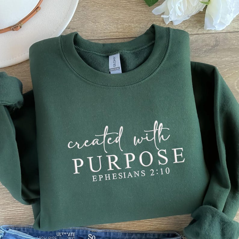 May include: A dark green sweatshirt featuring the text "created with PURPOSE" and "Ephesians 2:10" in white. The garment is folded on a wooden surface, with a white hat and flower visible in the background.