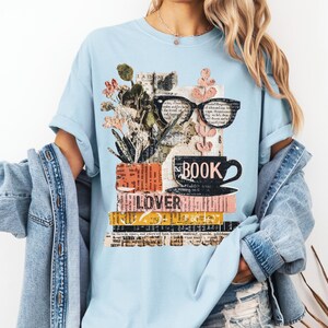 May include: Light blue t-shirt featuring a graphic design with books, eyeglasses, a coffee cup, and flowers. The word "BOOK" is printed on the cup, and "LOVER" is printed on the books. The design has a collage aesthetic.