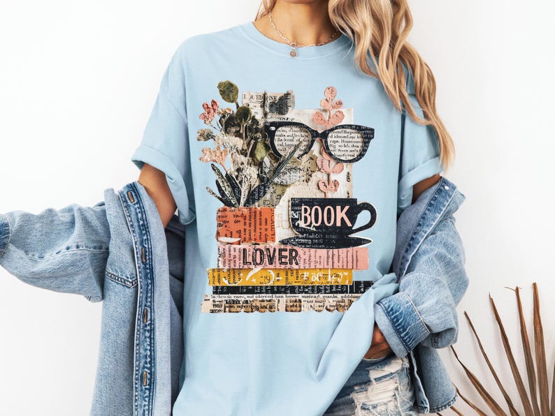 May include: Light blue t-shirt featuring a graphic design with books, eyeglasses, a coffee cup, and flowers. The word "BOOK" is printed on the cup, and "LOVER" is printed on the books. The design has a collage aesthetic.