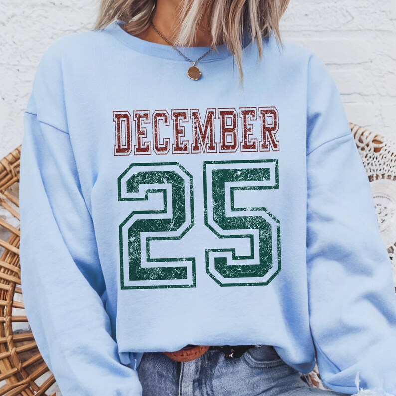 Pu&ograve; includere: Una felpa azzurra con la scritta "DECEMBER 25" in un carattere in stile collegiale usurato. La parola "DECEMBER" &egrave; rossa e il numero "25" &egrave; verde. Un capo casual e festivo.