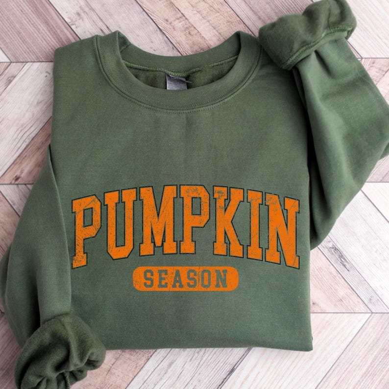 May include: An olive green sweatshirt with the words "PUMPKIN SEASON" printed in orange block letters. The sweatshirt is folded, with the sleeves and collar visible. The text has a distressed, vintage look.