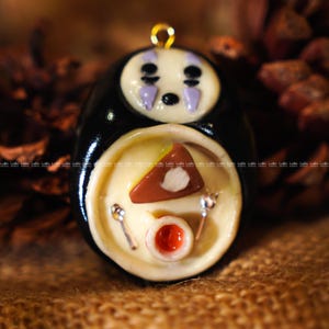 May include: A handmade pendant featuring a black, oval-shaped character with a white face and purple accents. The character is holding a plate with a slice of cake, a small bowl, and two tiny spoons. A gold-colored loop is at the top.