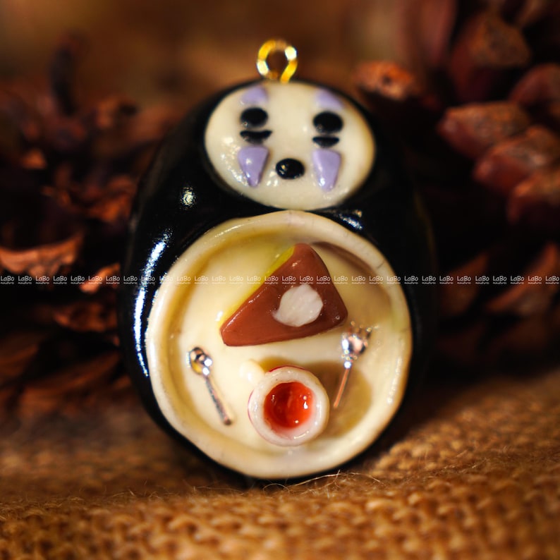 May include: A handmade pendant featuring a black, oval-shaped character with a white face and purple accents. The character is holding a plate with a slice of cake, a small bowl, and two tiny spoons. A gold-colored loop is at the top.