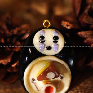 May include: A handcrafted pendant featuring a character from the animated film "Spirited Away." The pendant has a black body, a white face with purple markings, and a gold-colored loop. The lower section has a food-themed design.