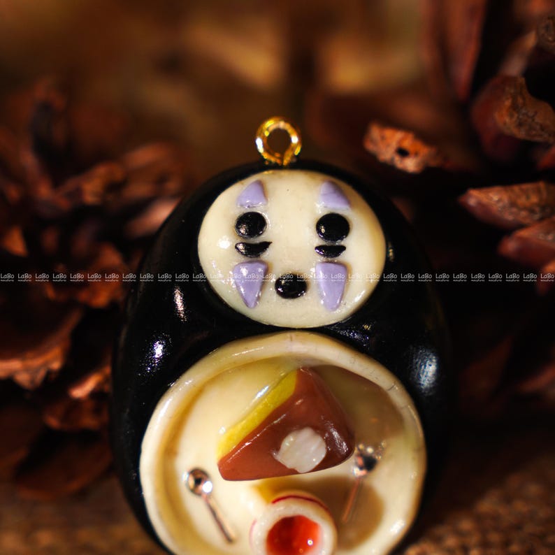 May include: A handcrafted pendant featuring a character from the animated film "Spirited Away." The pendant has a black body, a white face with purple markings, and a gold-colored loop. The lower section has a food-themed design.