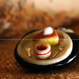 May include: Miniature dessert plate with a slice of cake topped with a white dollop, a small cup with a red stripe, and a tiny golden fork and spoon. The plate has a black rim and a light-colored center, creating a charming, detailed scene.