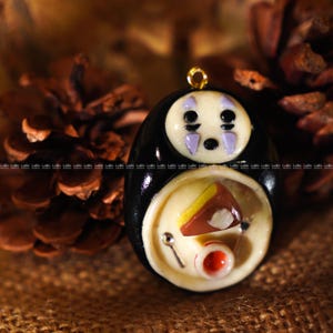 May include: A handmade pendant featuring a black figure with a white face and purple accents. The pendant has a gold-colored loop at the top. The lower portion of the pendant depicts a miniature meal with a slice of cake and a small cup.
