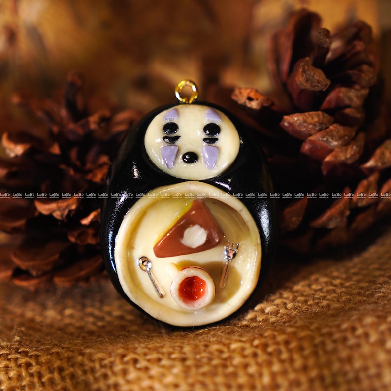 May include: A handcrafted pendant featuring a character from Spirited Away. The black and cream-colored figure has a gold loop for hanging. The lower portion depicts a plate with food, including a slice of cake and a small bowl.